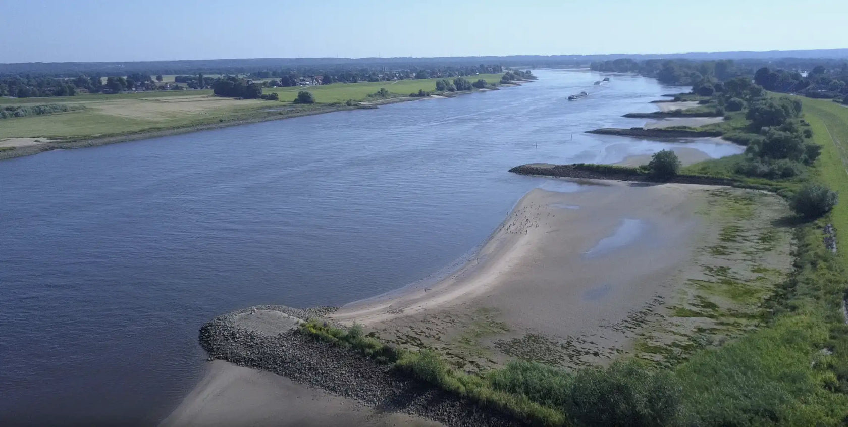 Ein breiter, ruhiger Fluss mit dunkelblauem Wasser fließt durch eine grüne Landschaft unter einem klaren Himmel.