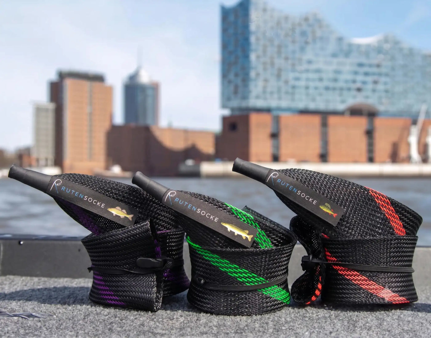 Drei schwarze geflochtene Halter für Angelnetz-Seil, jeweils mit einem unterschiedlich gefärbten Streifen, lila, grün und rot, und mit dem Branding „rutensocke“ sowie einem Fisch-Logo versehen.