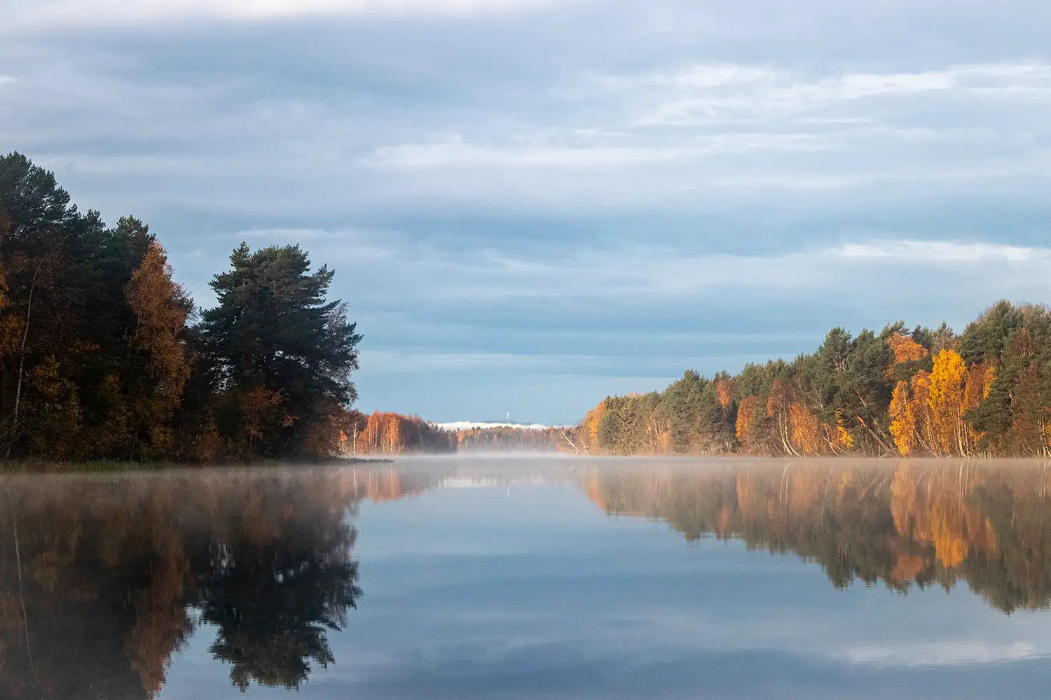 Ein ruhiger Herbstsee, dessen Oberfläche die lebendigen orangenen und grünen Bäume am Ufer unter einem sanften blauen Himmel widerspiegelt.