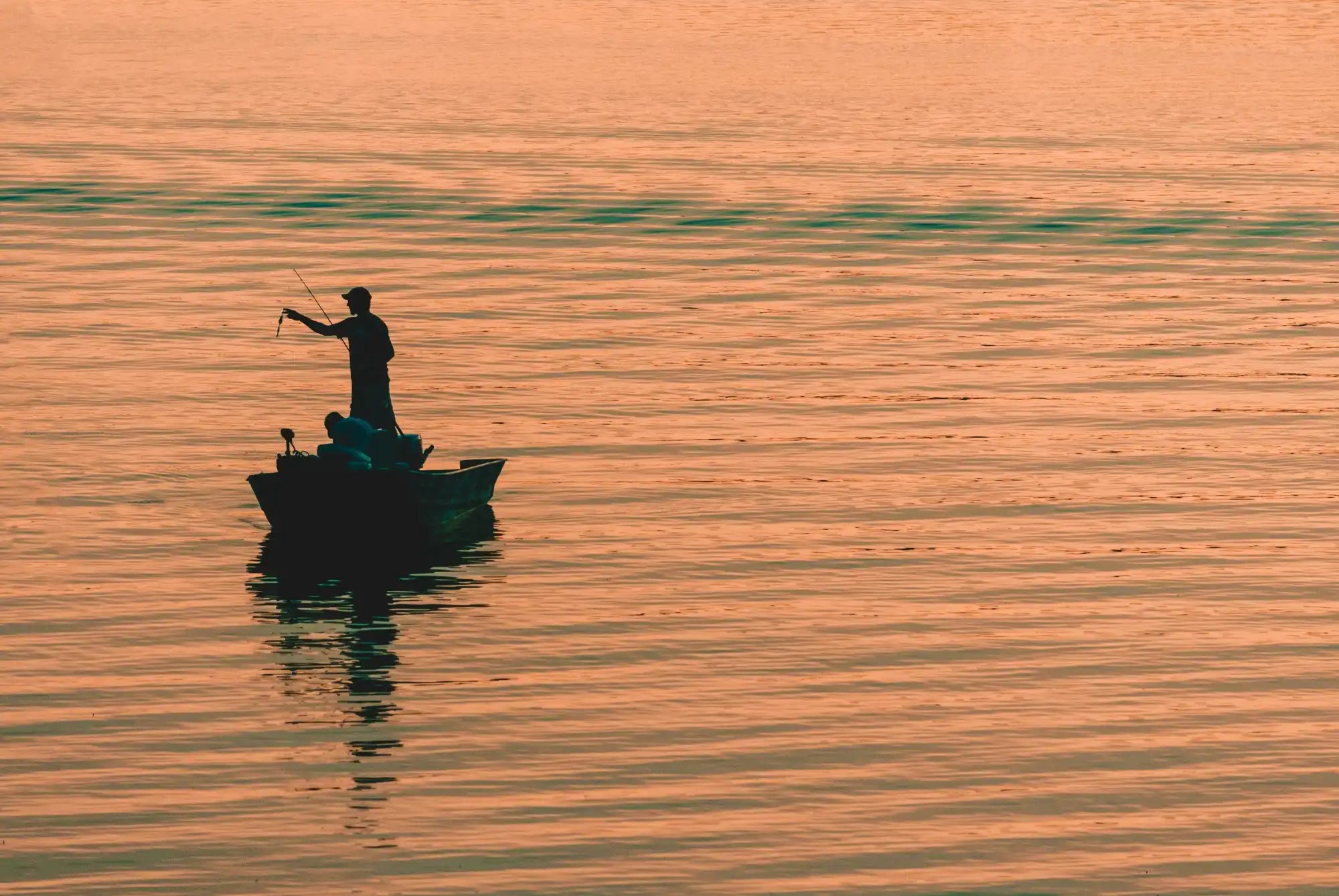 Silhouette einer Person, die bei Sonnenuntergang von einem kleinen Boot angelt.